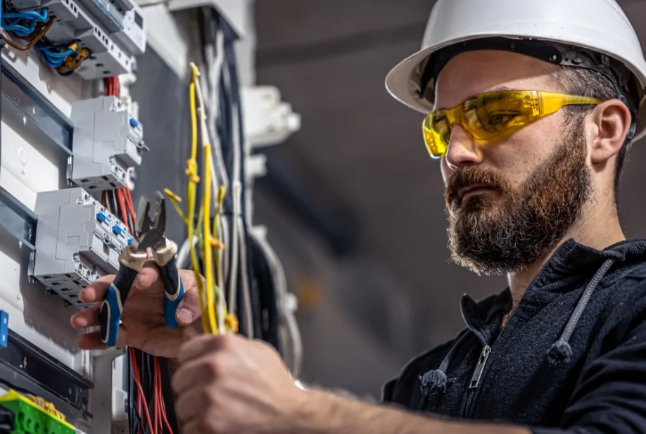 Eletricista Perto de Mim focado, com óculos de segurança, manuseando fios no painel.