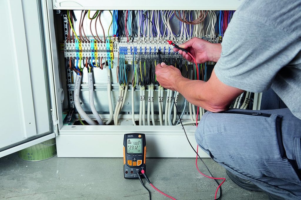 Eletricista 24 Horas analisando quadro de disjuntores com multímetro digital.